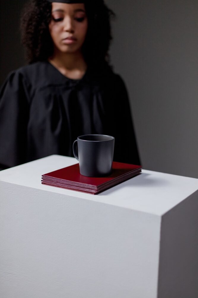 Universiteto absolventas su akademine mantija stovi šalia diplomų ir puodelio, simbolizuojančių aukštojo mokslo bei karjeros kelią.